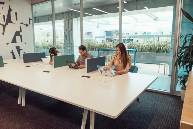 Trois personnes travaillent sur ordinateur portable autour d'une grande table blanche partagée, devant une baie vitrée, dans un espace de bureaux